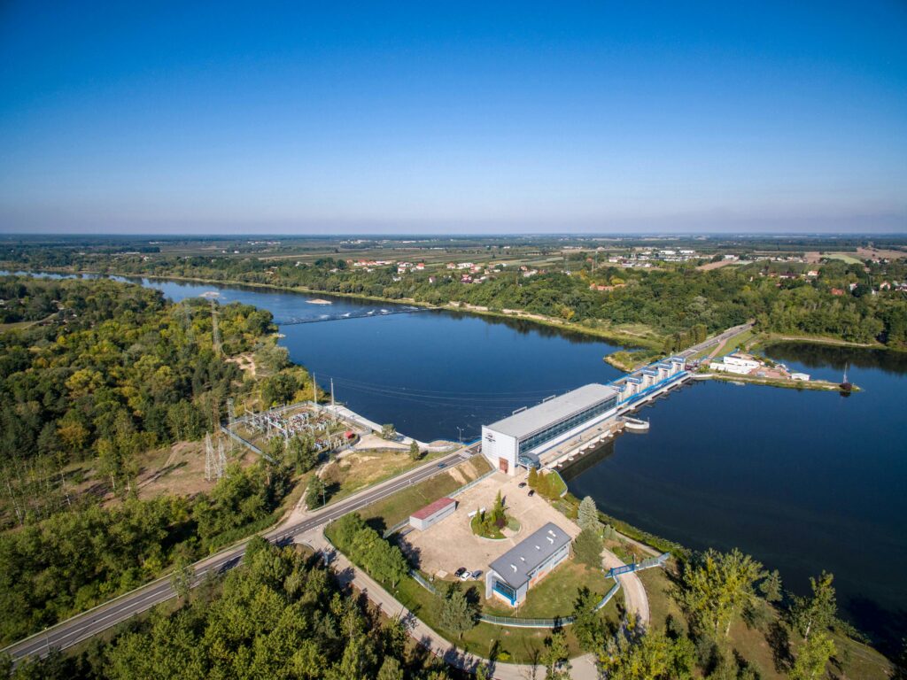 Home Scenic aerial view of a river dam surrounded by lush greenery under a clear sky.